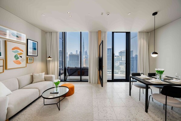 Una sala de estar moderna con sofá, mesa de centro y comedor con sillas, que cuenta con ventanas amplias con vista a la ciudad.