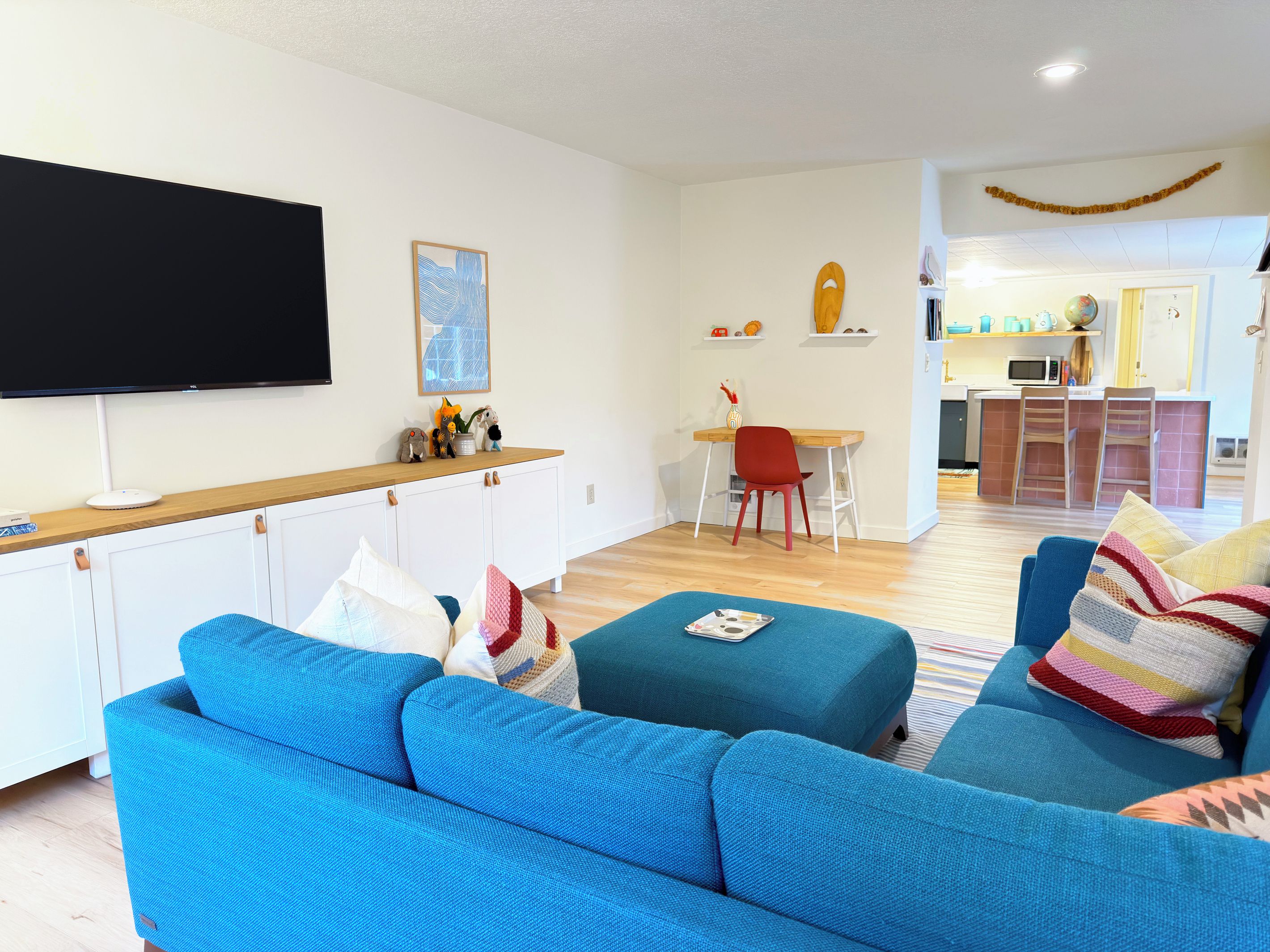 A modern living room with a flat-screen TV, a wooden dining table, and a kitchen area in the background.