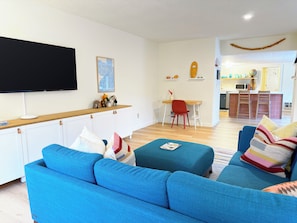 A modern living room with a flat-screen TV, a wooden dining table, and a kitchen area in the background. - 2 Bedroom Surfer's Bungalow in Seaside (Seaside)