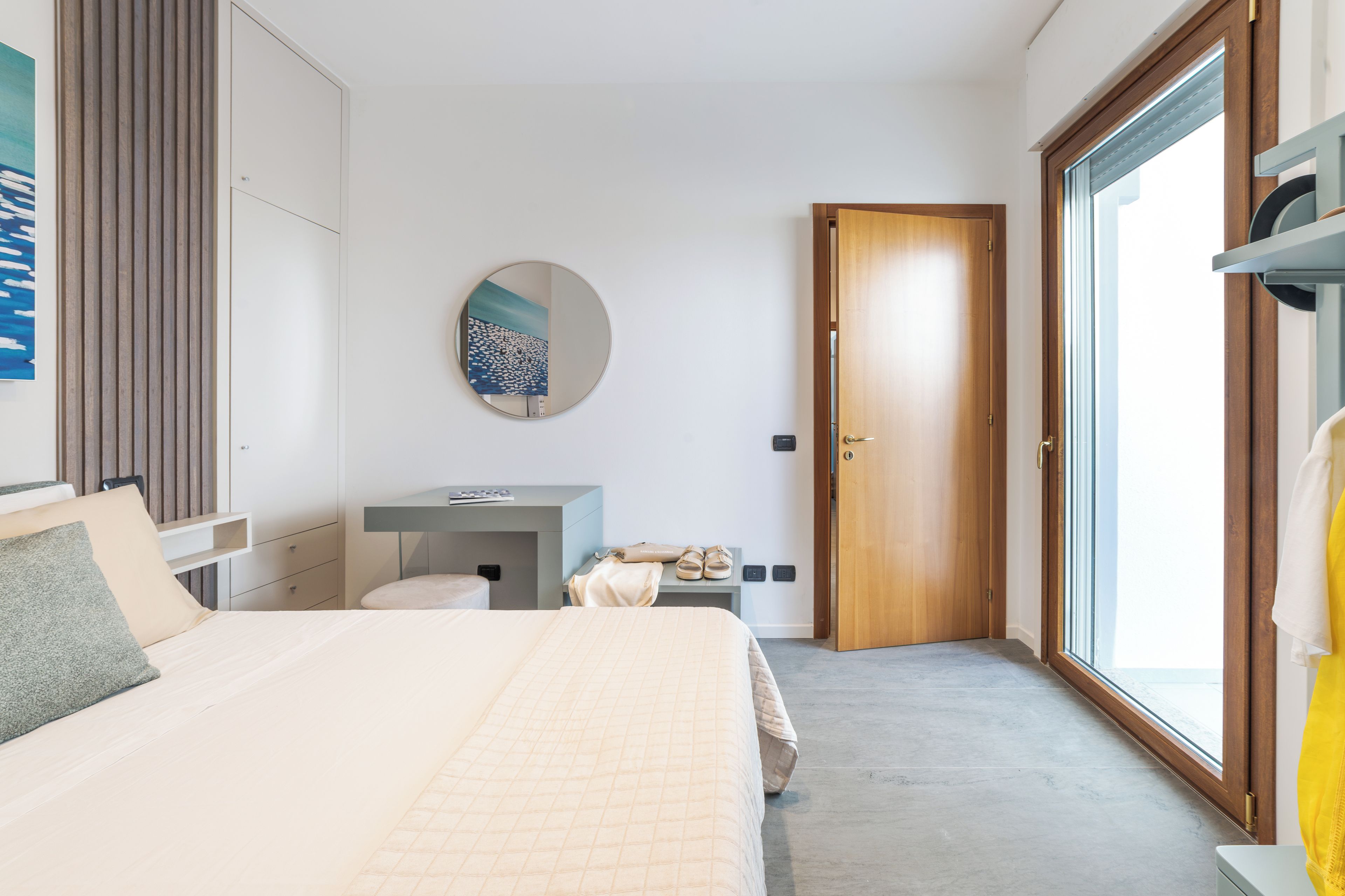 Une chambre d’hôtel moderne avec un grand lit, une table de chevet, un miroir, une armoire et une vue sur l’extérieur grâce à une porte vitrée.