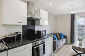 A modern kitchen with white cabinetry, a black countertop, and a grey sofa with blue cushions. - Modern Central Flat | Parking | Old Trafford (Greater Manchester)