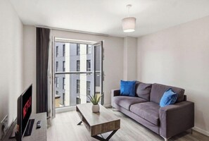 A modern living room with a grey sofa, a coffee table, a flat-screen TV, and a large window with a view of buildings. - Central Apartment | Parking | Old Trafford (Greater Manchester)
