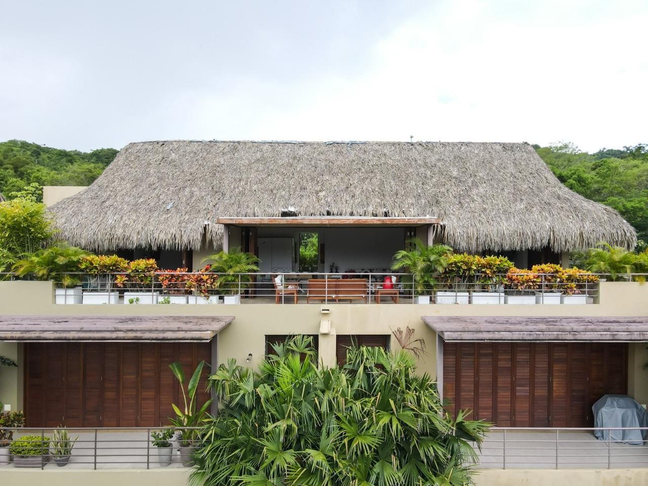 A two-story building with a thatched roof, a balcony with potted plants, and a view of a lush green landscape.
