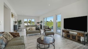 A spacious living room with a large flat-screen TV, comfortable seating, and a view of a green landscape through large windows. - Meadow View - outside Cheltenham with hot tub and games room, Sleeps 6 (Sandhurst)
