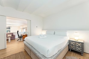 Quarto com cama, mesa de cabeceira, luminária e vista para uma sala de estar.
