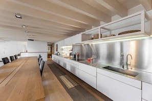 Cozinha moderna com longa mesa de madeira, armários brancos, eletrodomésticos de aço inoxidável e uma estante embutida com objetos decorativos.