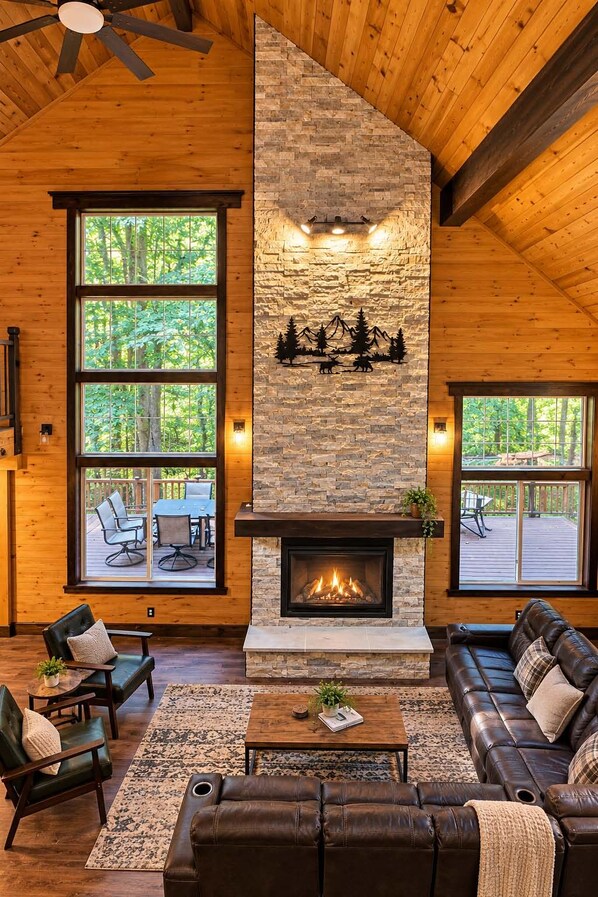 A cozy living room with a stone fireplace, leather furniture, and large windows overlooking a deck.