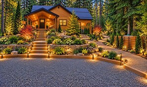 A log cabin with a stone staircase leading to the entrance, surrounded by a landscaped garden with various plants and trees.