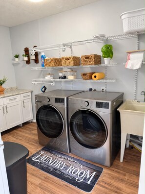 A laundry room with two front-loading washing machines, shelves above holding baskets and laundry supplies, and a doormat with a laundry-related message. - Spacious 5-bedroom house in wonderful Brandon with a wonderful Pool. (Brandon)