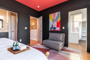 A modern hotel room with a bed, a grey sofa, a colorful abstract painting, and a wooden tray with bottles and towels. - Nashville-themed home with fire pit, sleeps 12, near 12 South & the Gulch (Nashville)