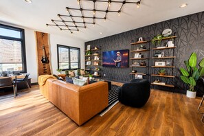 A modern living room with a wooden floor, a leather sofa, a black pouf, a TV, and a wall-mounted shelf with decorative items. - Fall in Nashville: skyline-view house, 4 ensuite baths, sleeps 12 (Nashville)