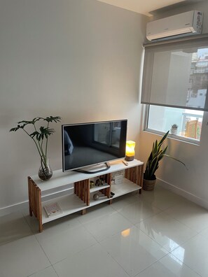 A modern living room with a flat-screen TV on a white stand, a potted plant, and a window with a view of buildings. - New Listing (Buenos Aires)