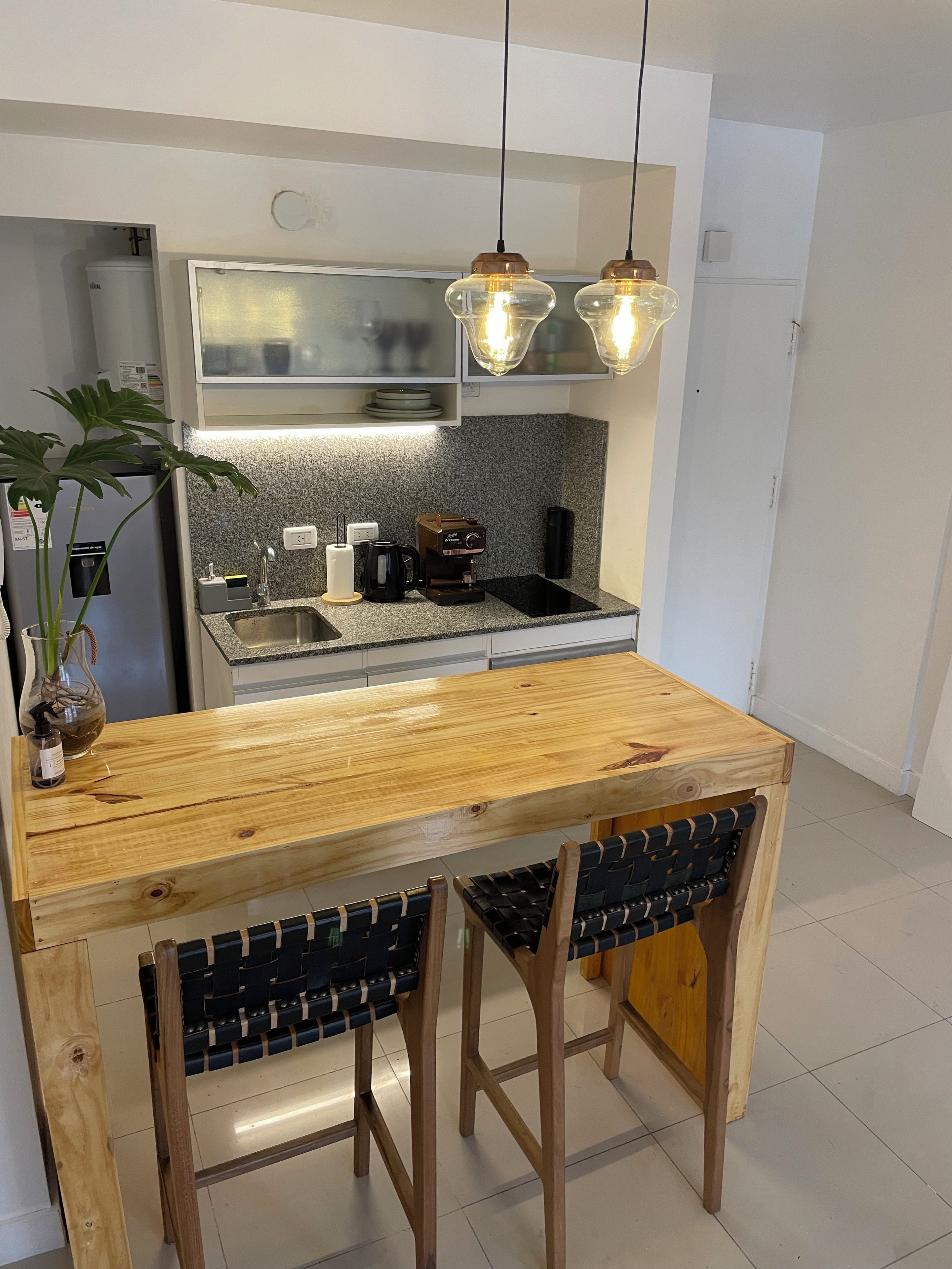 Una cocina con una mesa de madera y dos taburetes, una cafetera y una planta.