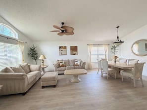 A spacious living room with a beige sofa set, a dining table with chairs, a ceiling fan, and a large mirror. - Greek Villa w/ Sunny Pool just 20 Min to Disney (Kissimmee)