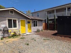 A two-story building with a yellow door, a covered outdoor seating area, and a gravel driveway. - Private, comfy, colorful 2 bed 2 bath home close to everything (Sacramento)