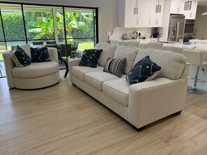 A modern living room with a beige sofa and armchair, a dining area with a glass table, and a kitchen with white cabinets and stainless steel appliances. - Lovely newly remodeled 3-bedroom, 3 bath house in charming Bonita Springs (Bonita Springs)