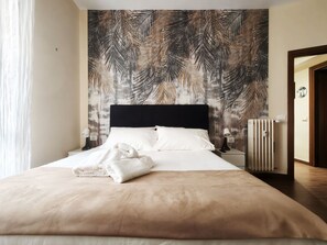 A hotel room with a bed, white linens, a black headboard, a wall with a palm leaf pattern, a bedside lamp, and a radiator. - Jireh vacation home - Two Bedroom Apartment, Sleeps 5 (Rome)