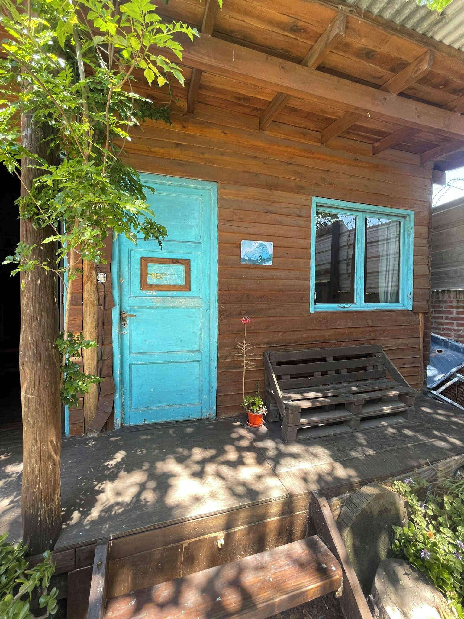 Una cabaña de madera con una puerta azul, una ventana con cortina y un banco afuera.