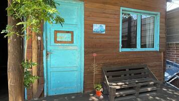 Una cabaña de madera con una puerta azul, una ventana con cortina y un banco afuera.