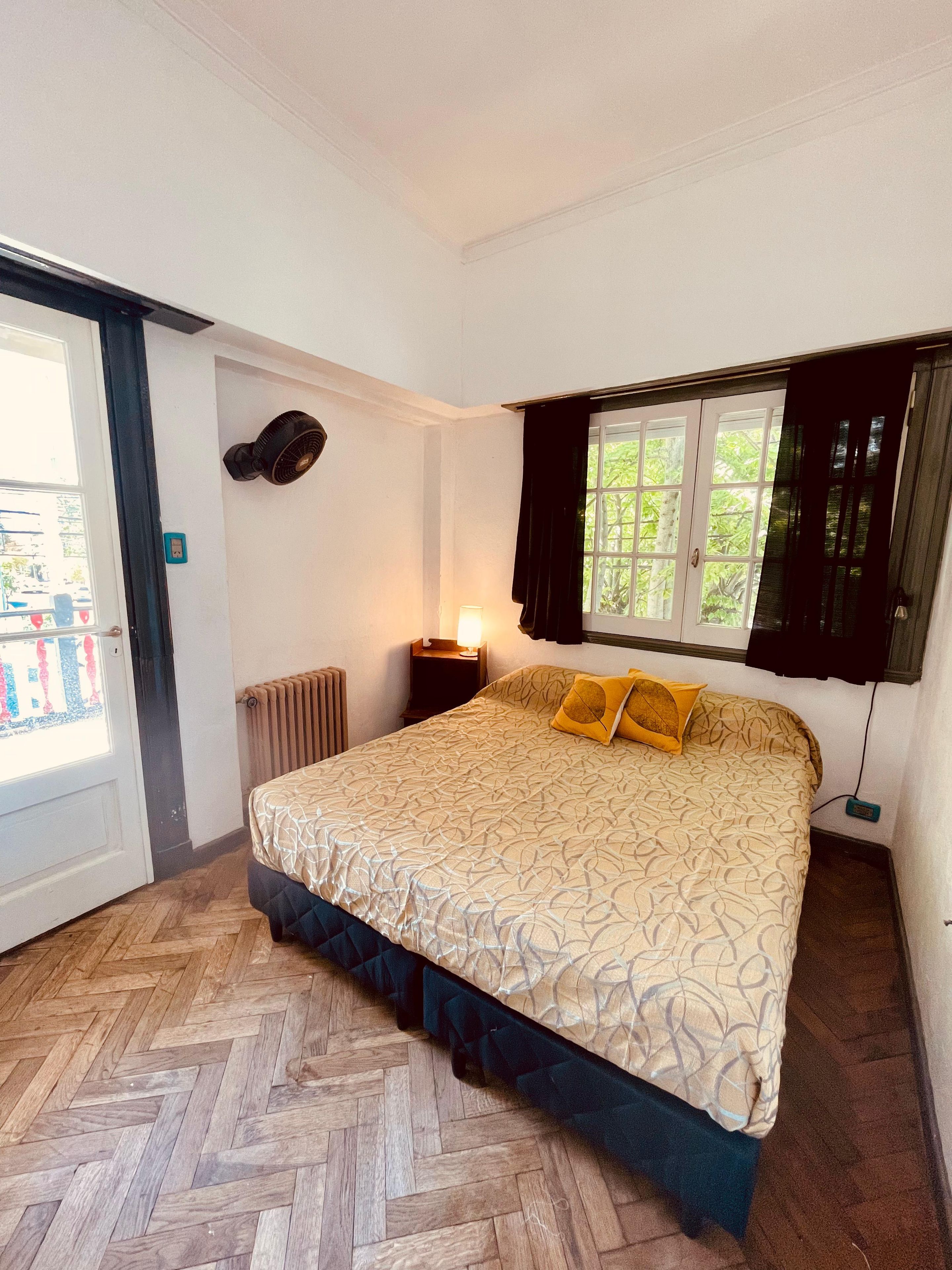A bedroom with a bed, a standing fan, a window with curtains, a radiator, and a door.
