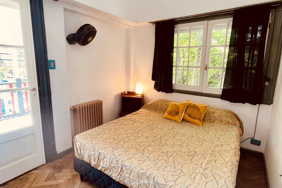 A bedroom with a bed, a standing fan, a window with curtains, a radiator, and a door.