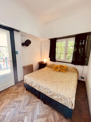 A bedroom with a bed, a standing fan, a window with curtains, a radiator, and a door.