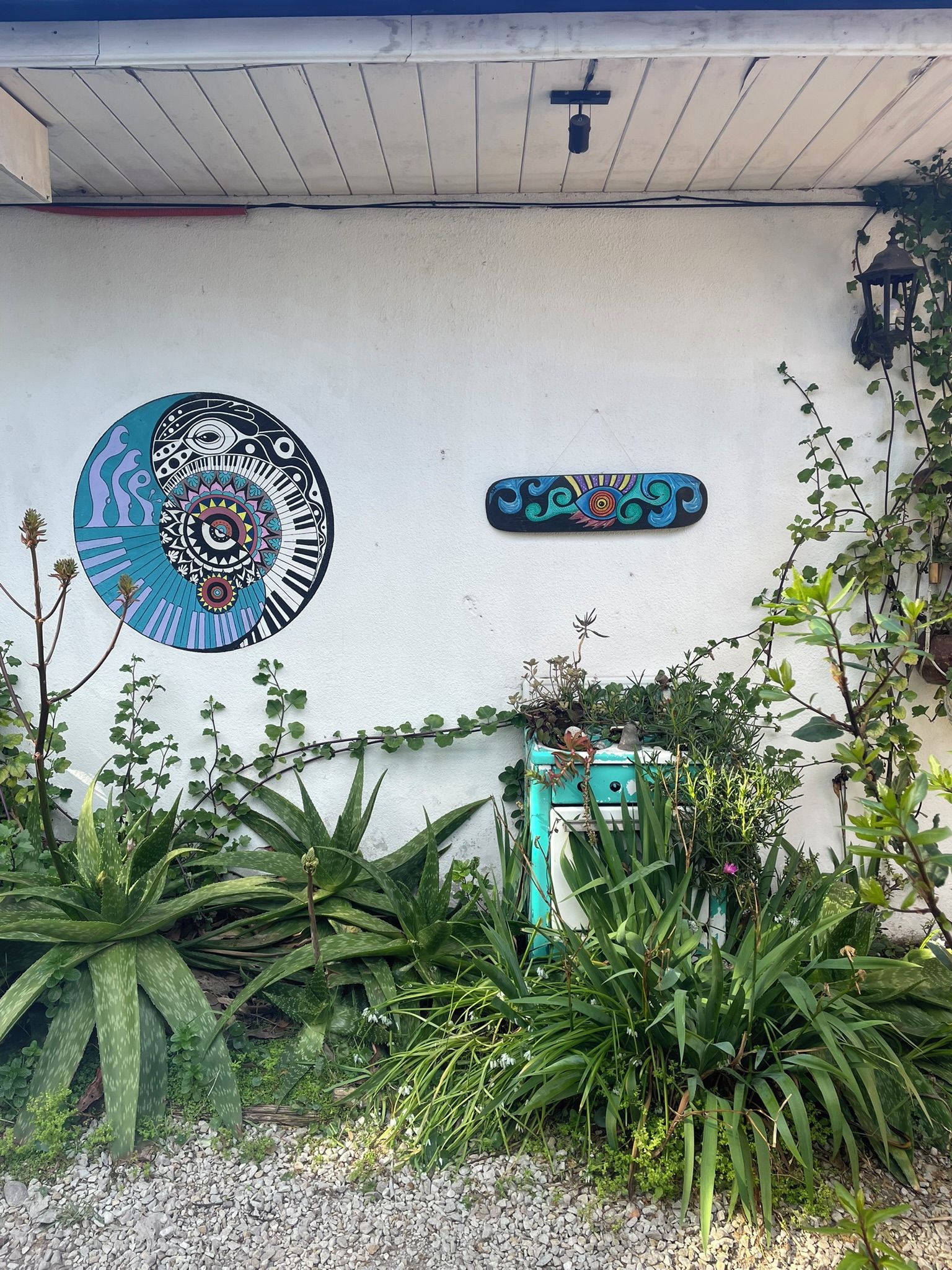 Paredes com um mural circular e uma placa retangular, rodeadas por vegetação e uma pequena construção.