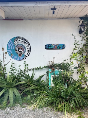 Una pared con un mural circular y un letrero rectangular, rodeada de follaje y una pequeña construcción.