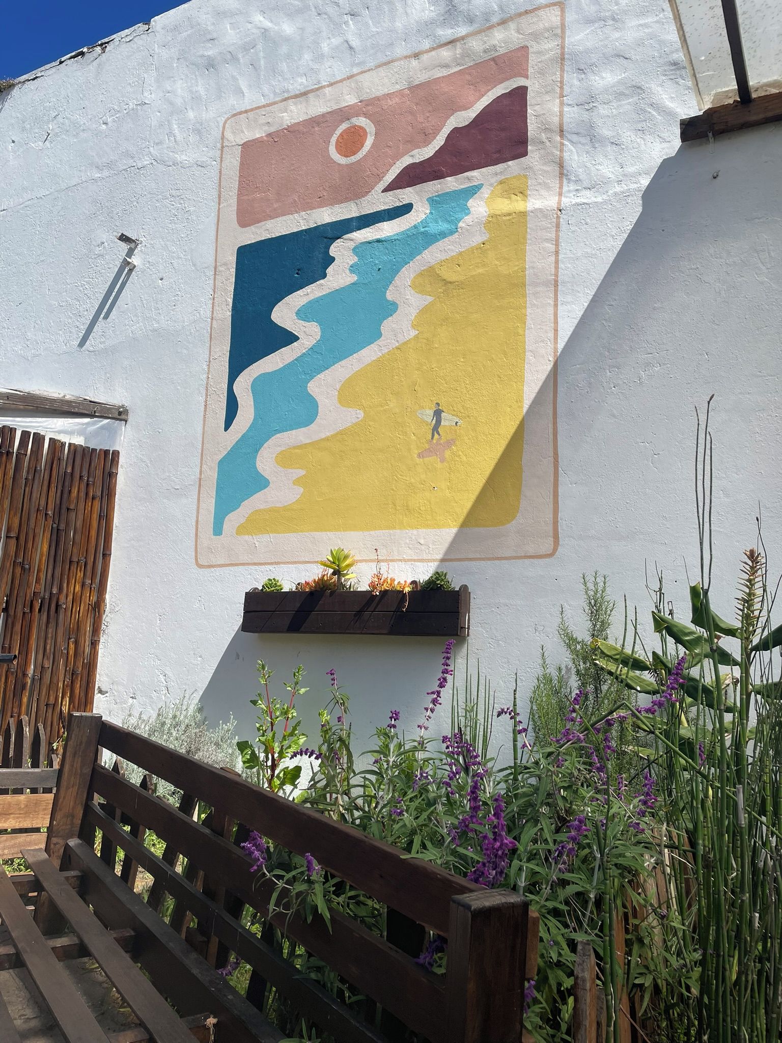 A wall with a colorful mural, a wooden bench, and various plants.