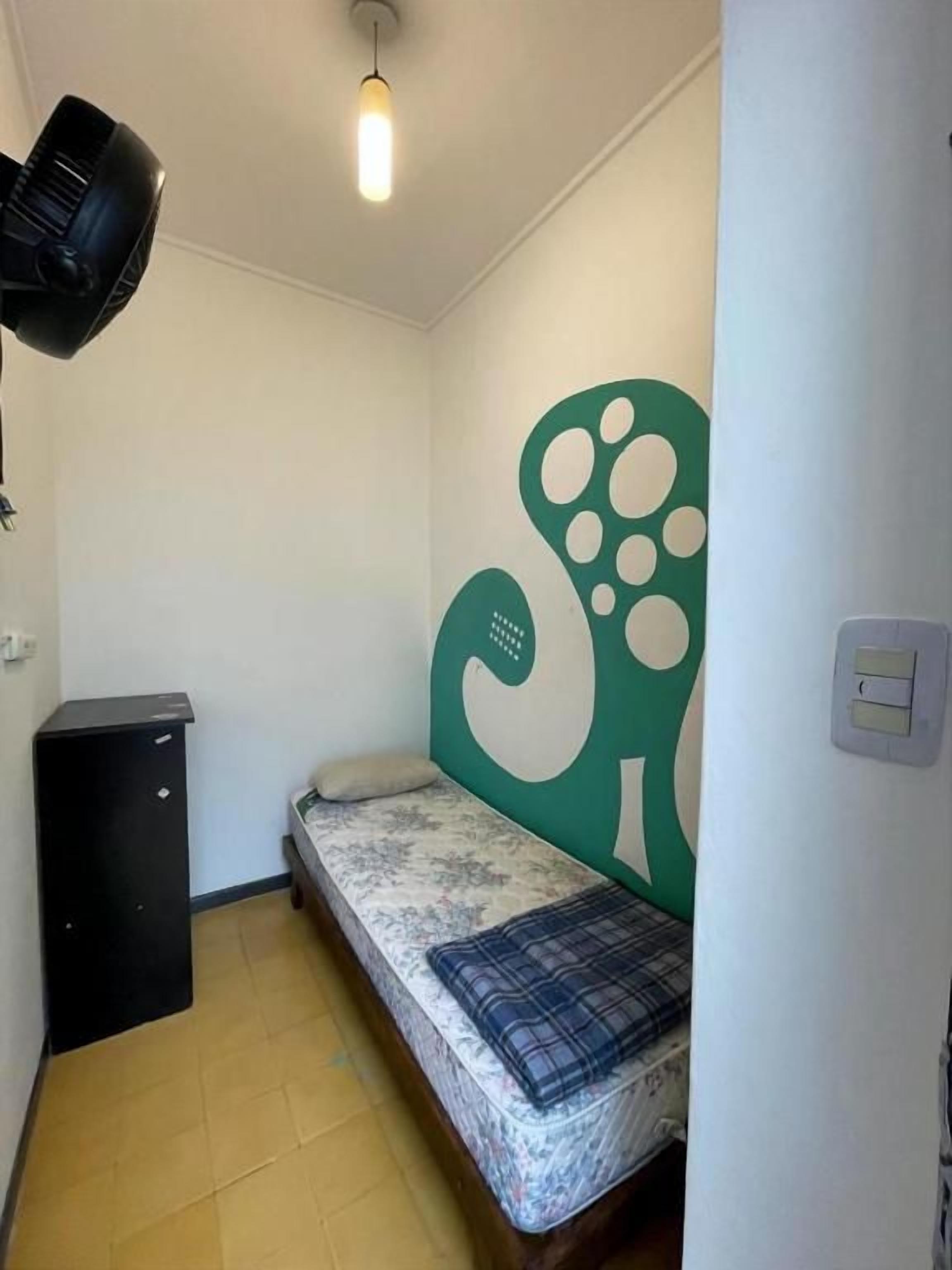 Quarto pequeno com cama, luminária na parede e um ornamento verde na parede.