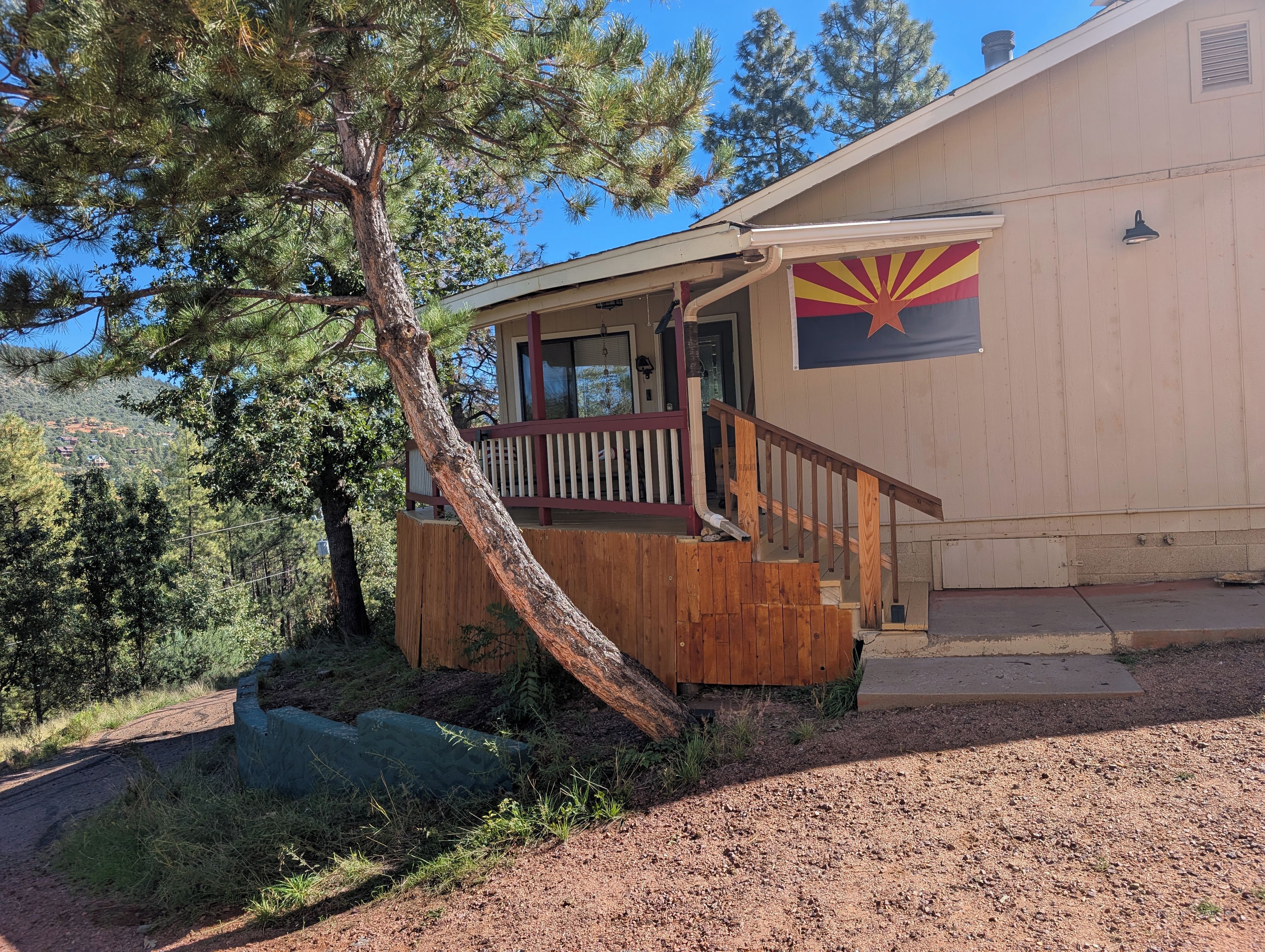 A building with a wooden staircase leading to a porch, a flag on the side, and a tree in front.