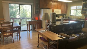A living room with a wooden floor, a black leather sofa, a wooden coffee table, and a dining area with a wooden table and chairs.