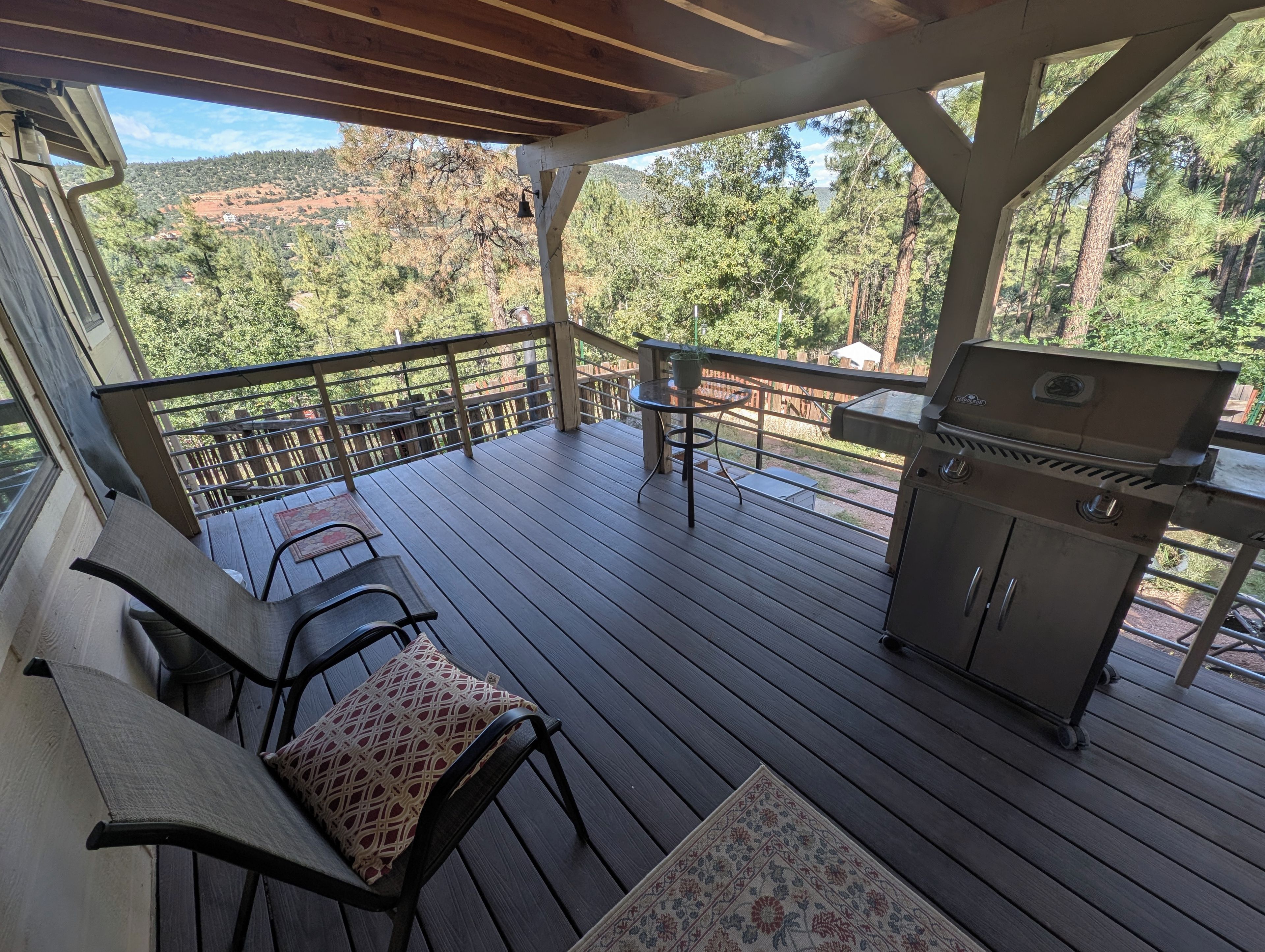 A covered outdoor deck with a grill, seating area, and a view of a forested landscape.