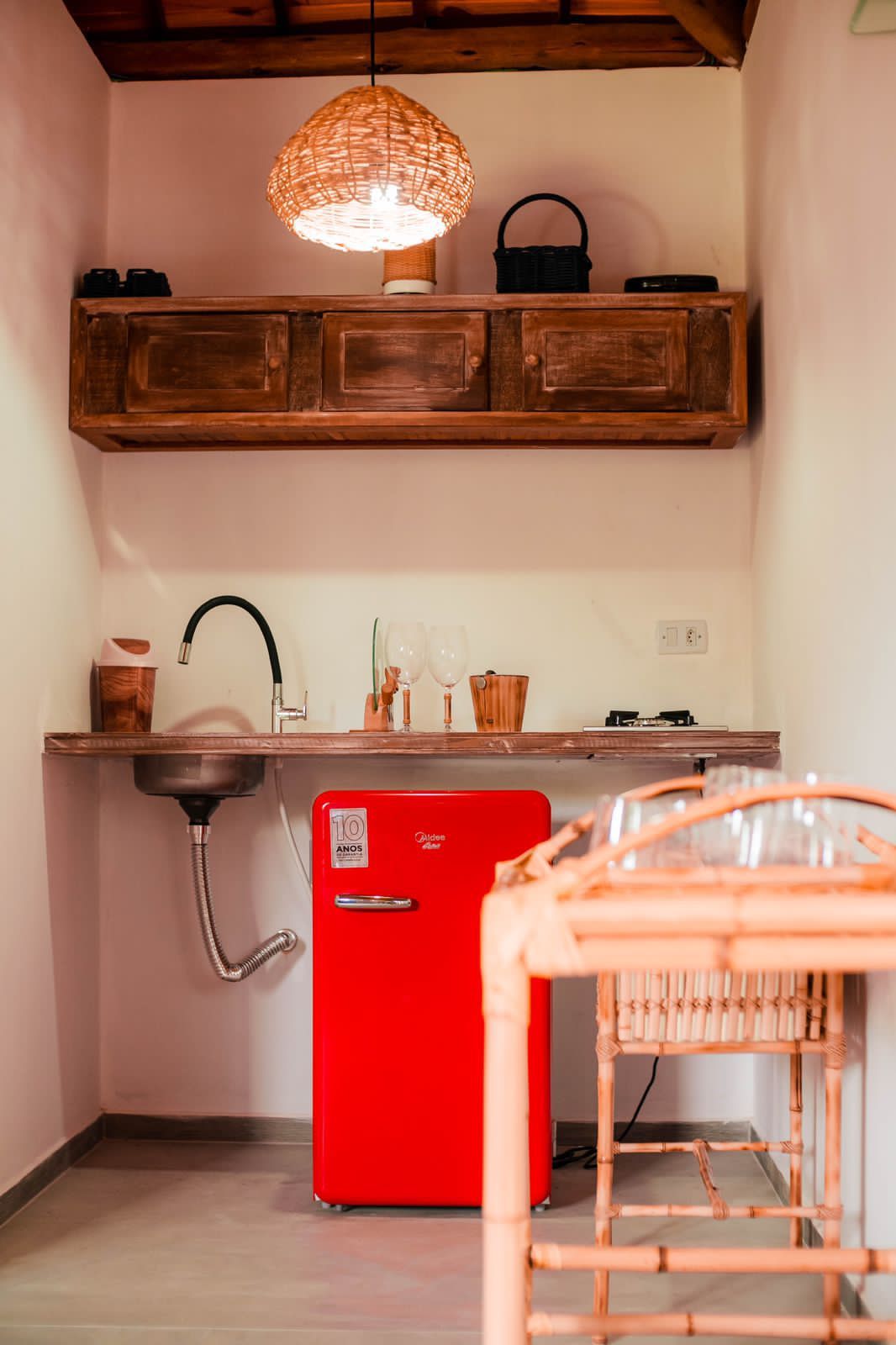 Une cuisine avec un réfrigérateur rouge, des meubles de rangement en bois, un luminaire suspendu et une chaise en bambou.