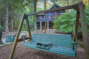 A wooden swing set with a bench in a forested area, surrounded by trees and a cabin with large windows. - Mountaintown Creek Lodge | Ellijay, GA (Gilmer County)