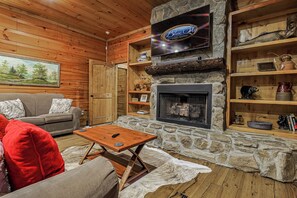 A cabin interior with a stone fireplace, wooden walls, a television mounted above it, and a sofa with red cushions. - Mountaintown Creek Lodge | Ellijay, GA (Gilmer County)