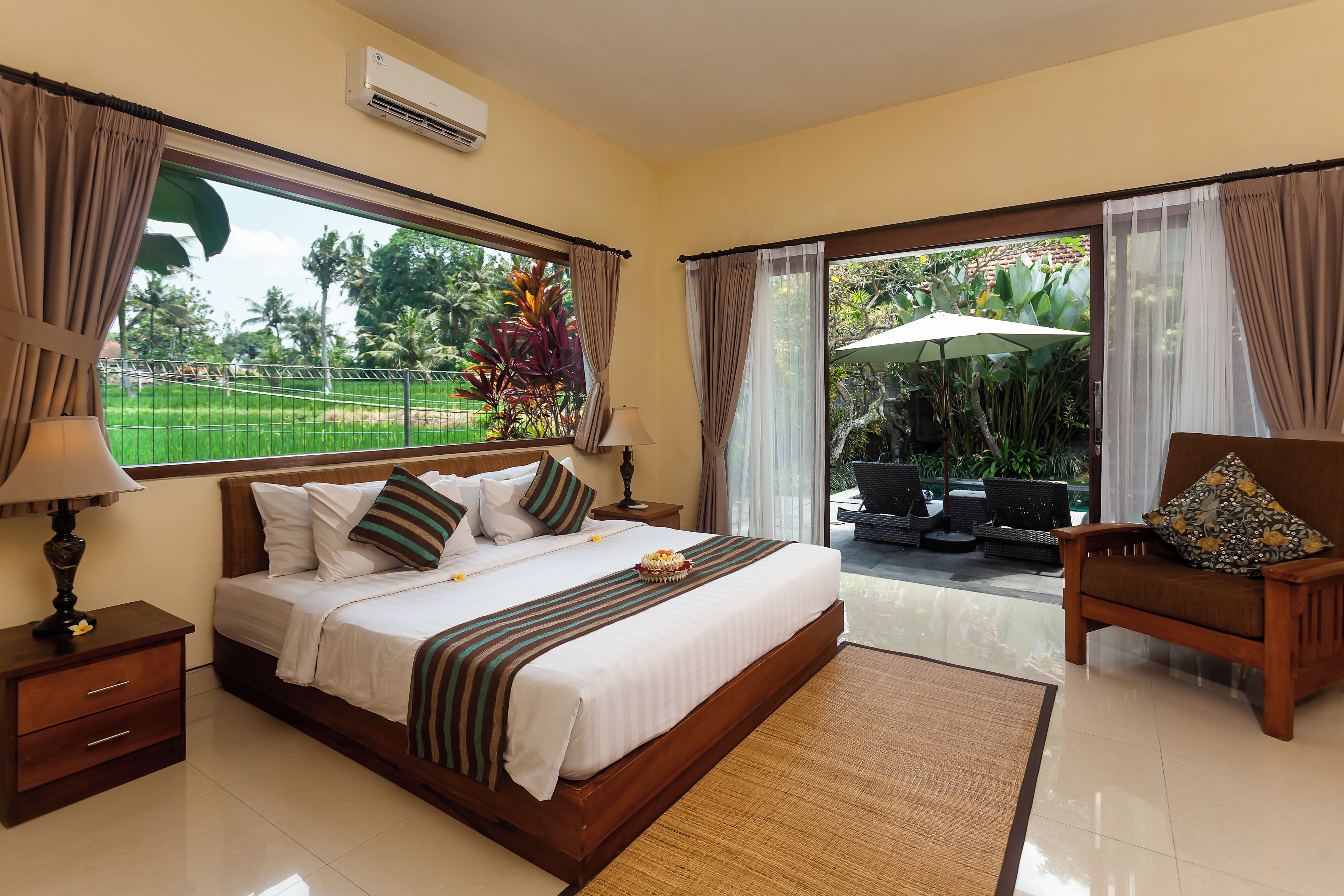 A hotel room with a large bed, a nightstand, a lamp, a view of a garden through a sliding door, and a chair with a floral pillow.