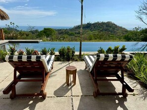 Duas cadeiras de descanso com almofadas listradas voltadas para uma piscina e a vista de uma colina arborizada.