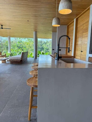 A modern kitchen with a long countertop, wooden stools, pendant lighting, and a view of greenery outside. - NEW 6BR Contemporary Villa Near Santa Teresa • Ocean Views • Saltwater Pool (Santa Teresa)