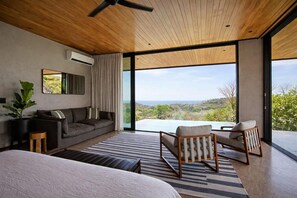 A modern bedroom with a large window, a ceiling fan, a sofa, a chair, and a bed. - NEW 6BR Contemporary Villa Near Santa Teresa • Ocean Views • Saltwater Pool (Santa Teresa)