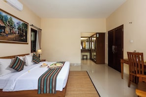 A hotel room with a large bed, a dining table, a chair, a painting on the wall, and a view into the corridor. - Kadiga Villas - 2 BR pool Villa (Bali)