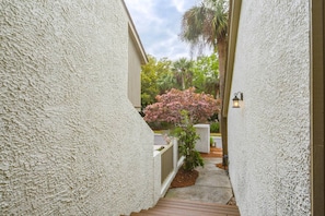 A narrow pathway flanked by textured walls, leading to a gated area with a small tree and a pink flowering bush. - Elegant Sea Pines Villa Steps From The Beach (Hilton Head Island)