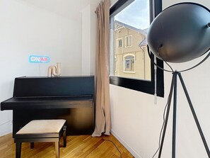 A room with a piano, a stool, a window with a view of buildings, and a floor lamp. - Mercury · Mercury House 110 m² | Private parking | Train station 500 m away (Montreux)