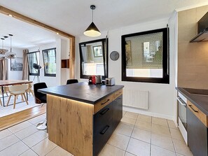 A modern kitchen with a central island, wooden cabinetry, and a dining area with a table and chairs. - Mercury · Mercury House 110 m² | Private parking | Train station 500 m away (Montreux)