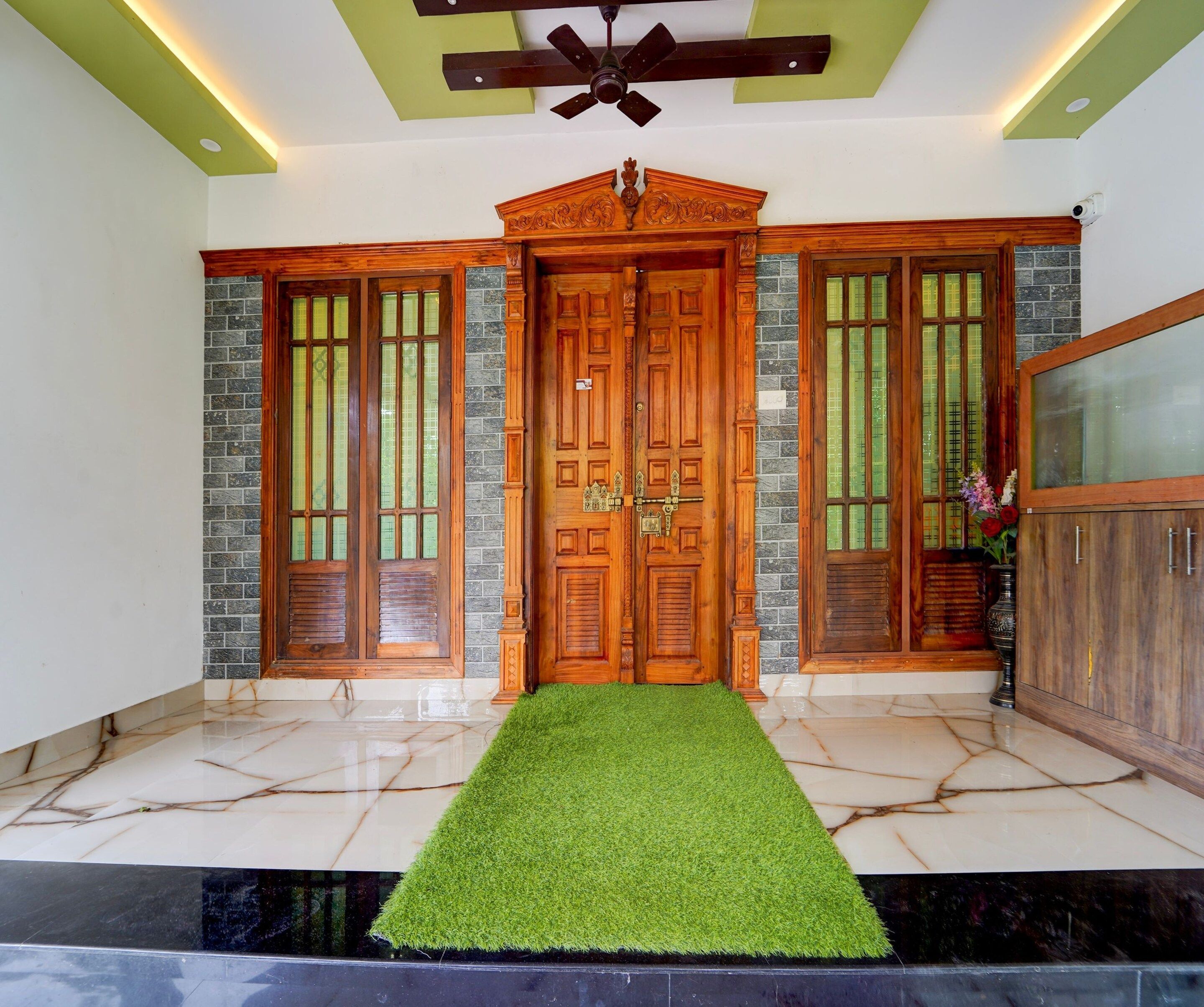 A foyer with a wooden double door, a green artificial grass entrance, a ceiling fan, and a tiled floor.