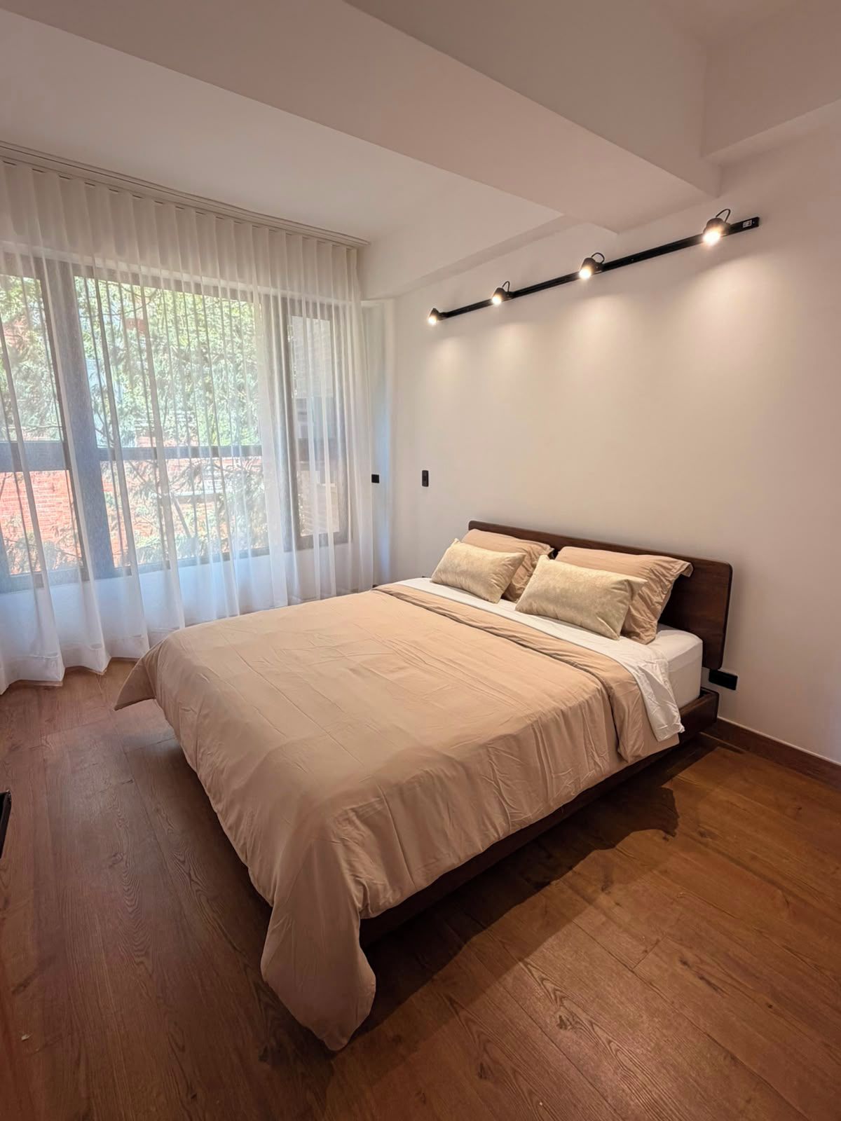 A modern bedroom with a large bed, wooden flooring, and a window with sheer curtains.