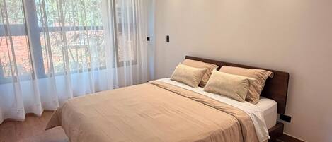 A modern bedroom with a large bed, wooden flooring, and a window with sheer curtains.
