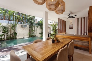 A modern dining area with a wooden table, chairs, and a view of an outdoor swimming pool. - The Arca Villa - 2 BR Pool Villa (Bali)