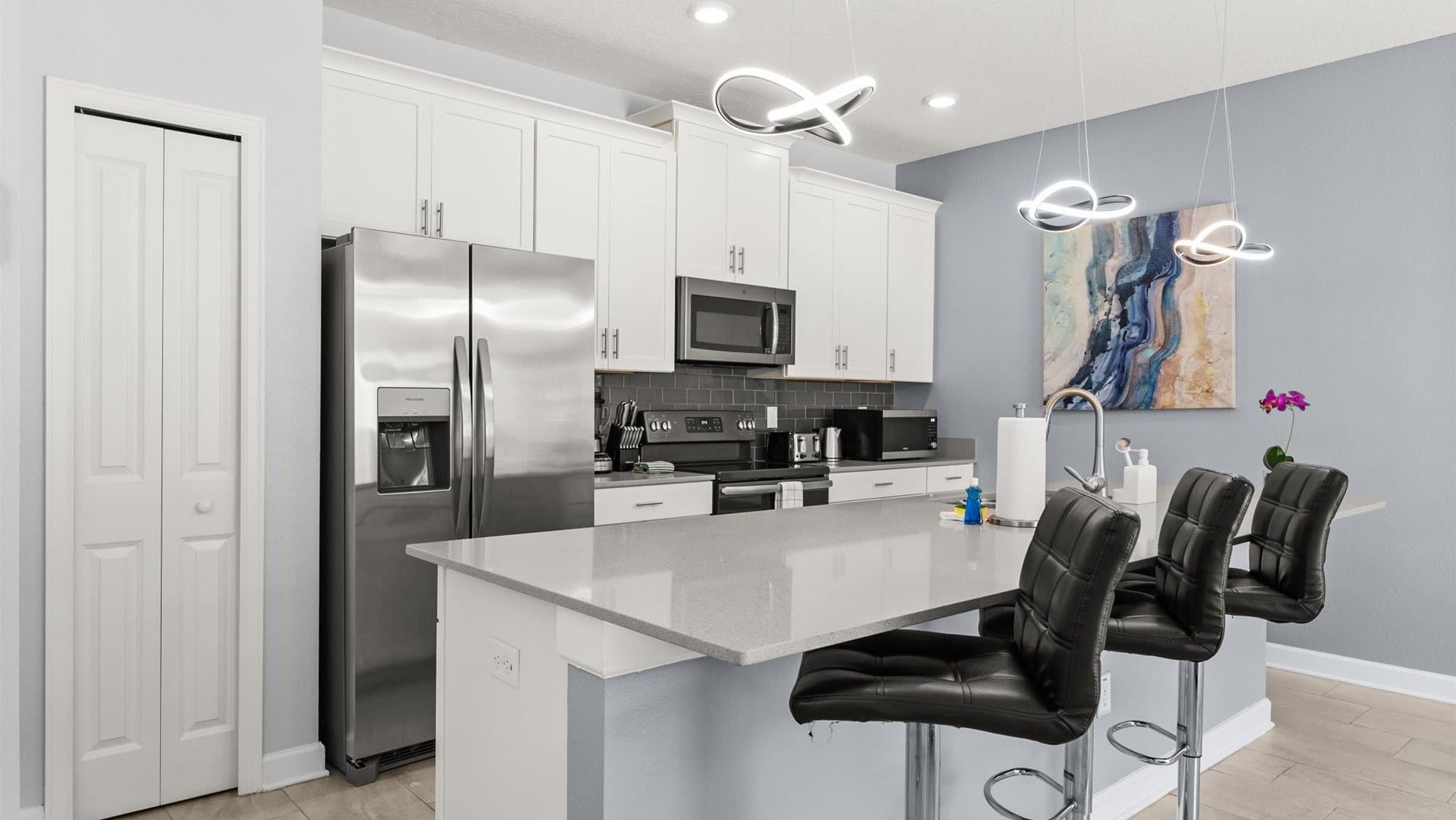 Una cocina moderna con armarios blancos, electrodomésticos de acero inoxidable, una isla central con taburetes y una lámpara de diseño contemporáneo.