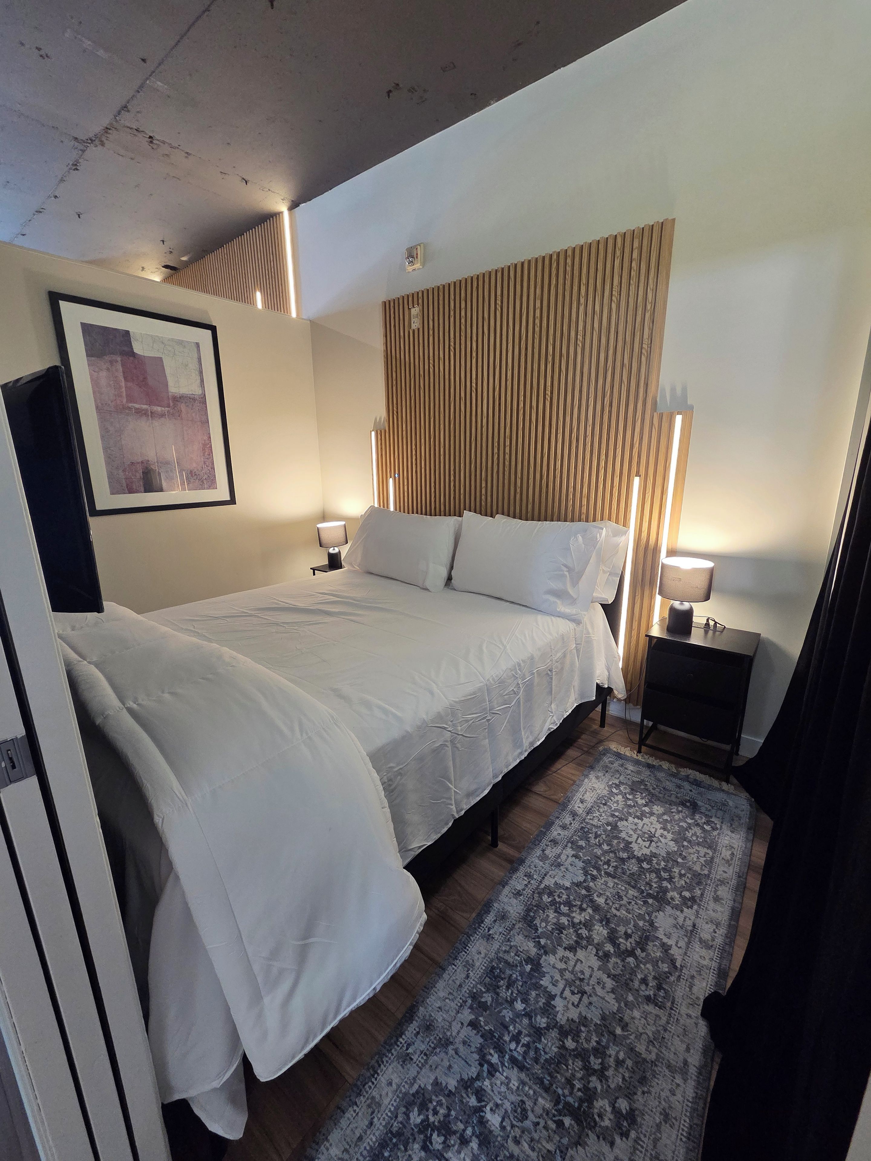 Ein Hotelzimmer mit Bett, Nachttischlampen, Teppich, Holz-Kopfteil, gerahmtem Bild an der Wand und einem kleinen Nachtkästchen.